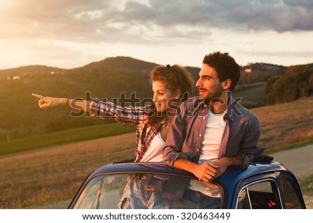 A loving couple, on a summer afternoon, watching sunset from the roof of an old car, around the classical landscape of Tuscany, vineyards and farmland. Woman points to look at a point to her boyfriend