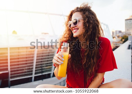 Beautiful young woman in sunglasses holds a bottle with fresh juice, looking away, sitting on marble slab in a sunny day, outdoors. Foto d'archivio © 