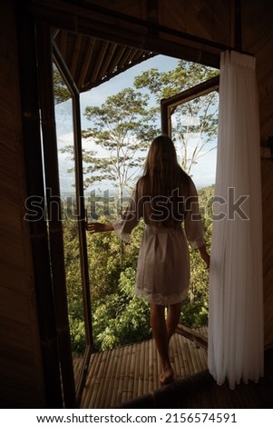 Slender girl in a bathrobe goes outside through a window in a bamboo house. Outside the window is a beautiful view of the tropical forest. 商業照片 © 
