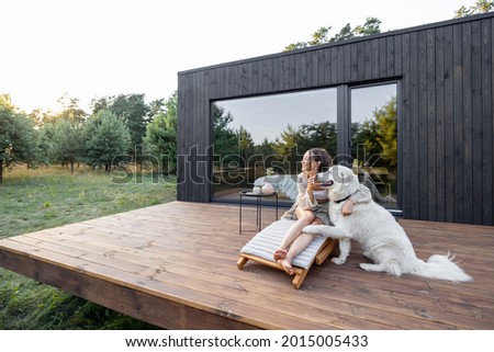 Woman enjoys the nature while sits on sunbed on wooden terrace near the modern house with panoramic windows near pine forest while hugs her pet. Concept of solitude and recreation on nature