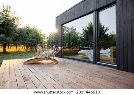Young woman resting on sunbed and reading on a tablet on the wooden terrace near the modern house with panoramic windows near pine forest. Concept of solitude and recreation on nature Foto stock © 
