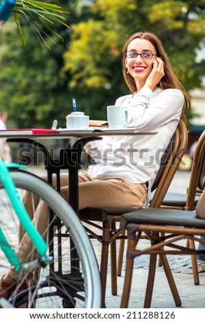 Young woman sitting in the open air cafe with cup of coffee and bicycle and speaking phone in the morning in the city centre