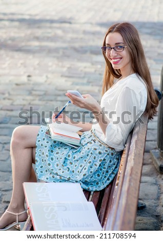 Young woman sitting and studying on the bench with books typing message on the phone in the city