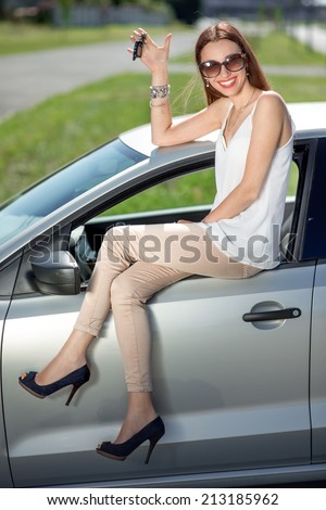 Young woman holding keys to new car smiling at camera and sitting on the car door