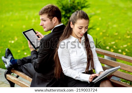 Young Business couple sitting on the bench and reading or working with tablets outdoors.
