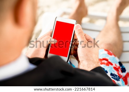 Businessman holding cellphone with empty screen sitting on the sunbed dressed in suit and shorts on the beach