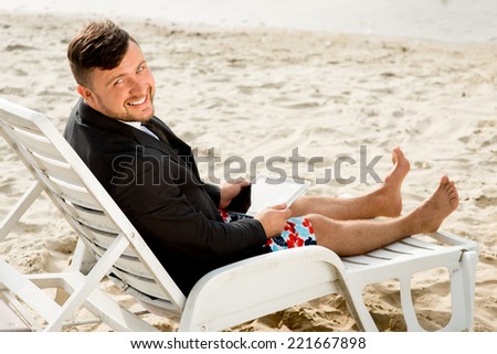 Businessman dressed in suit and shorts working with laptop on the sunbed at the beach