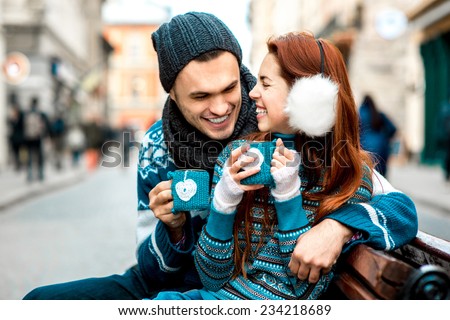 Young loving couple dressed in blue sweater sitting with knitted coffee cups on the bench at the old city in winter