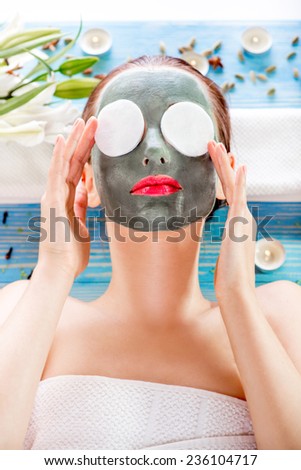 Young woman with spa facial mask on her face and cotton tampons on her eyes lying on blue table with flower, candles and sea salt in the beaty salon