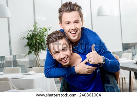 Friendly brothers twins having fun riding piggyback in the white home or restaurant interior. Family relationship between two brothers