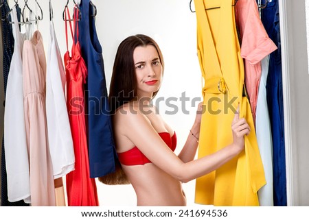 Young upset woman in underwear choosing dress to wear in the wardrobe