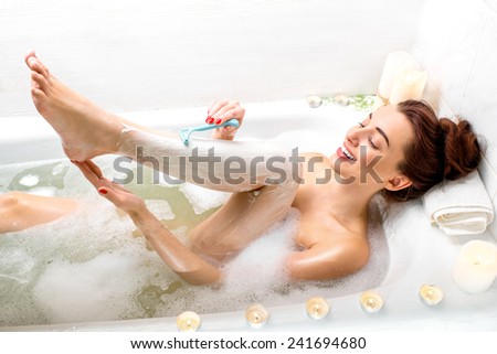 Young and positive woman shaving her legs in the bath with foam and candles.