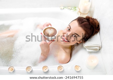 Young woman enjoying coffee while lying in the bath with foam and candles