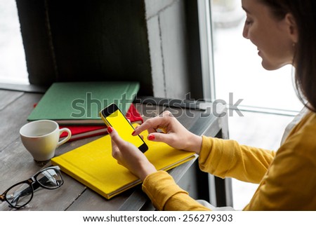 Woman using smart phone on the table with colorful books in the cafe near the window. Phone with empty screen for your application