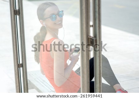 Young sport woman with phone waiting at the bus station. View through the glass reflection