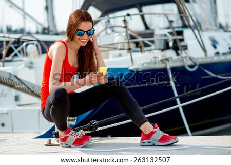 Young sport woman using mobile sitting near the yachts at the marine