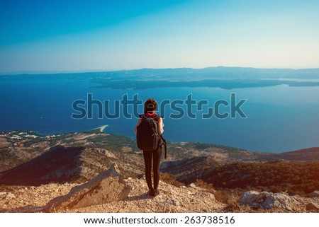 Young woman traveler with backpack standing on the top of mountain. General back view