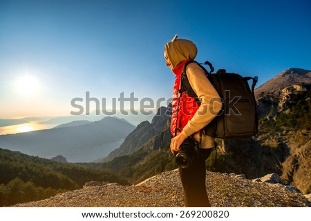 Young traveler photographer with photo camera and backpack standing on the top of mountain and looking on the sunset