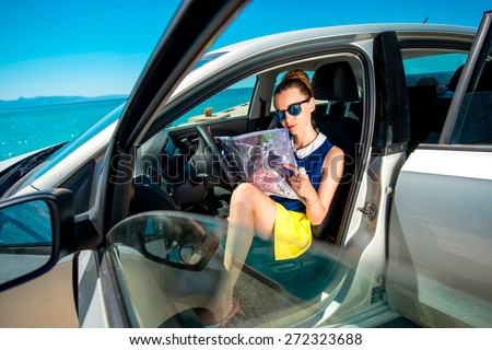 Young woman traveler sitting with paper map in the car and looking where to go on blue sea background