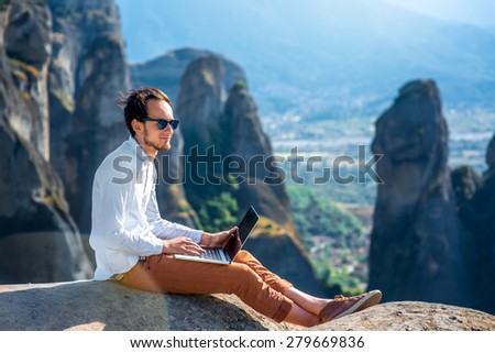 Well-dressed man working with laptop sitting on the rocky mountain on beautiful scenic clif background near Meteora monasteries in Greece.