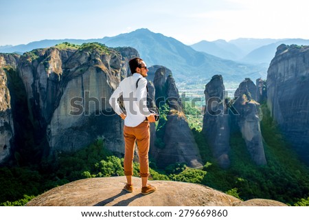 Professional well-dressed photographer standing on the top of mountain on beautiful scenic clif background near Meteora monasteries in Greece. Back view, general plan.