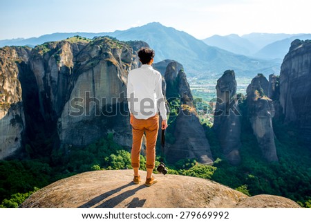 Professional well-dressed photographer standing on the top of mountain on beautiful scenic clif background near Meteora monasteries in Greece. Back view, general plan.