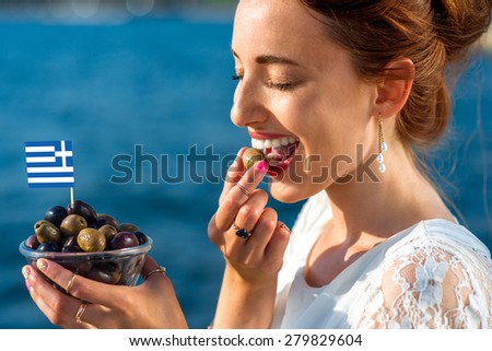 Smiling woman eating fresh green and black olives with greek flag outdoors on blue sea background