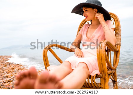 Young woman in pink dress and big black hat resting in the wicker chair on the beach in cloudy weather.