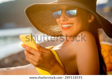 Young woman in swimsuit, hat and sunglasses using yellow mobile phone on the beach at sunset. Communication by phone on the summer vacation.
