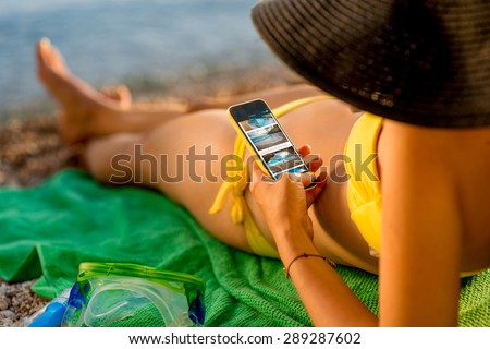 Young woman in swimsuit watching website with beach photos on mobile phone lying on the green towel on the beach. Focused on the hand with phone.