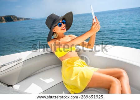 Young woman in yellow swimsuit and skirt taking selfie with digital tablet on the yacht floating in the sea
