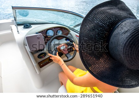 Young and pretty woman in yellow skirt and swimsuit with hat and sunglasses driving luxury yacht in the sea. Top view without face focused on hands and helm