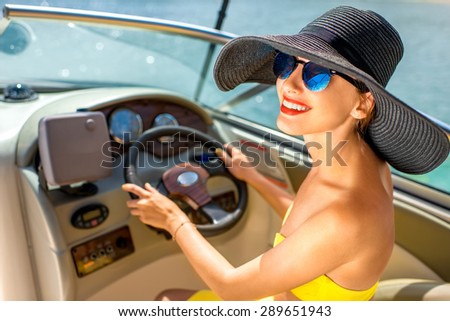 Young and pretty woman in yellow skirt and swimsuit with hat and sunglasses driving luxury yacht in the sea.