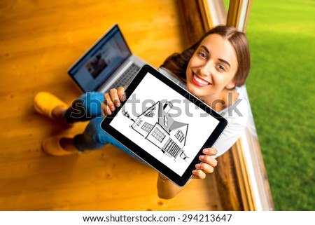 Young and cute woman showing digital tablet with house drawing sitting near the window in cozy wooden cottage. Projecting cottage concept