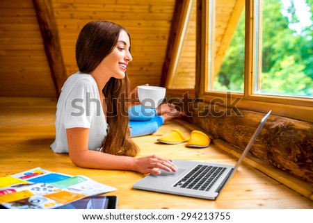 Young woman in jeans and white shirt lying on the floor with laptop, tablet and coffee cup near the window in cozy wooden cottage