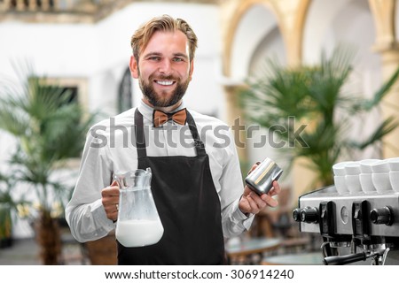 Smiling barista in uniform holding milk pitcher and jar filled with milk standing near the coffee machine in the cafe