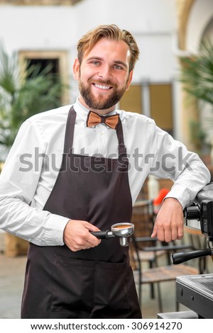 Handsome bearded barista in uniform standing with coffee handle near the coffee machine in the classical cafe patio