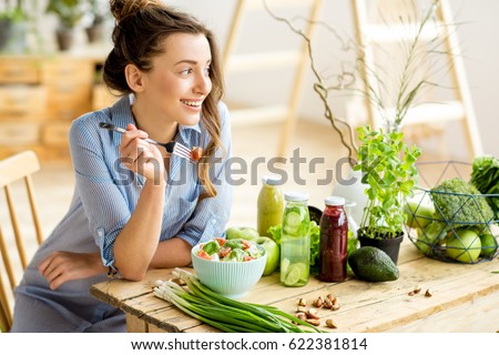 Young and happy woman eating healthy salad sitting on the table with green fresh ingredients indoors Foto d'archivio © 