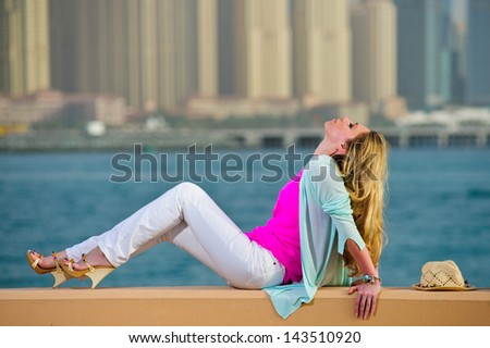 Yang European woman poses in stone fence in front of seaside modern city.