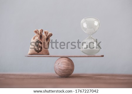 Write sand clock or hourglass and dollar bagson a balance scale in equal position on wood table. Financial concept : Time value of money, asset growth over time, depicts investment in long-term equity