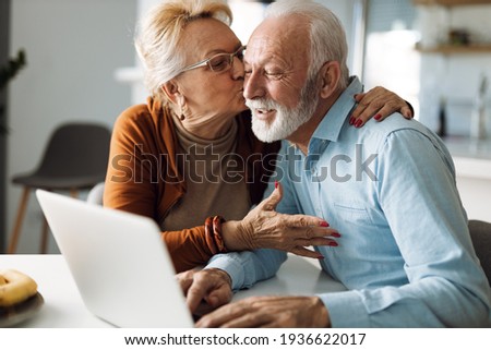 Mature woman kissing her mature husband while he uses a computer at home