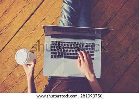 Laptop and paper cup of coffee in girls hands sitting on a wooden floor