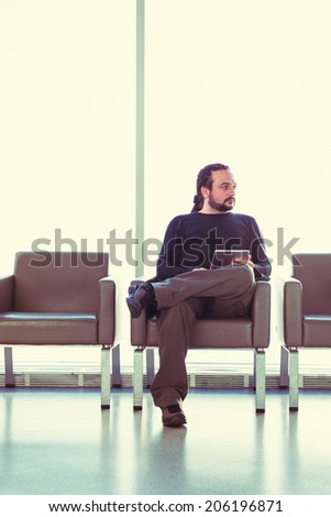 Handsome young man with dreadlocks using his digital tablet pc at an airport lounge, modern waiting room, with backlight.