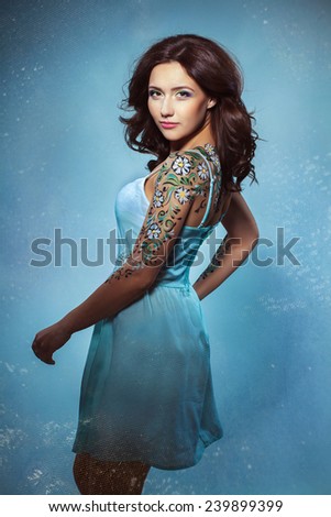 girl in a blue dress with a tattoo on a blue background. Beauty woman.Jewelry and beauty. perfect skin, brunette. Girl with a stylish haircut. Skin Care.