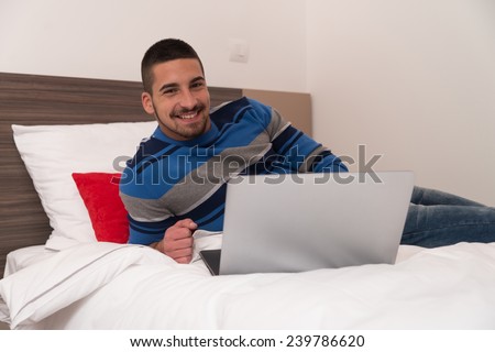 Young Male Student Lying On Bed And Having Fun With Laptop In Bedroom