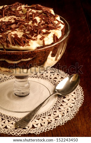 Chocolate sponge cake with vanilla cream and cherries in a big bowl on lacy napkin, silver spoon and rustic oak table