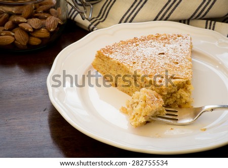 Homemade sweet lemon cake with almonds and oat flour, glass jar with almonds, slice with fork on white plate, old wood table