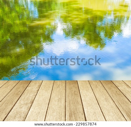 Empty perspective wood over the reflection of green trees and blue sky on water, product display template