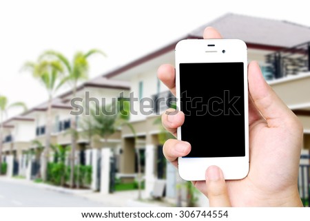Hand holding white mobile smart phone over blurred house background, smart home concept, template, mock up