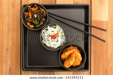 Asian vegetarian dish composed with three black bowls with shrimps, rice noodles, fried vegetables and chinese chopsticks on a square plate. Composition on a old styled wooden table.